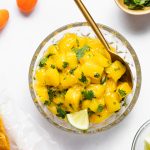 Mango habanero salsa in a bowl with a spoon, surrounded by cilantro, habanero peppers, limes, and chips.