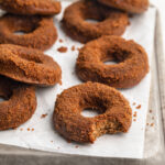 healthy vegan apple cider donuts on a sheet of parchment paper, one with a bite taken out.