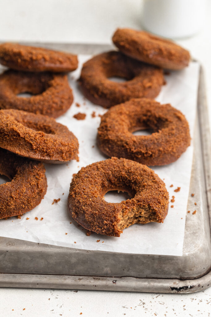 healthy vegan apple cider donuts on a sheet of parchment paper, one with a bite taken out.