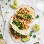 overhead image of three instant pot shredded chicken tacos on a table with fresh cilantro and limes on the side