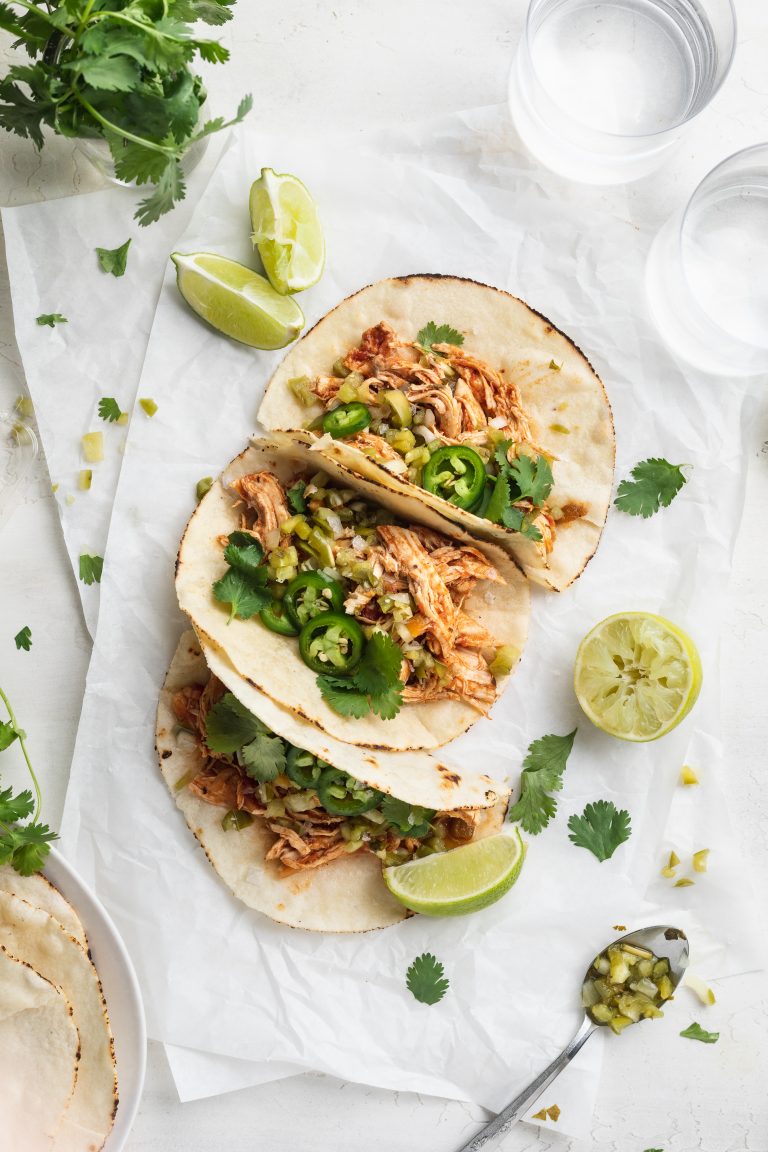 overhead image of three instant pot shredded chicken tacos on a table with fresh cilantro and limes on the side