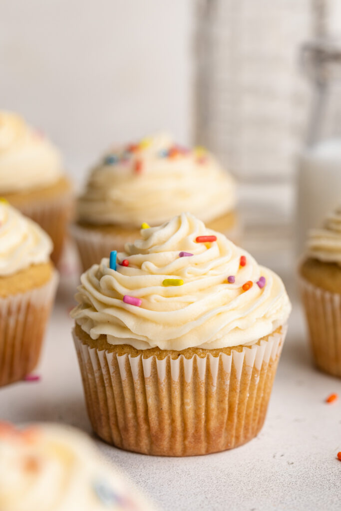 close up image of a gluten-free vanilla cupcake with vanilla frosting and rainbow sprinkles.