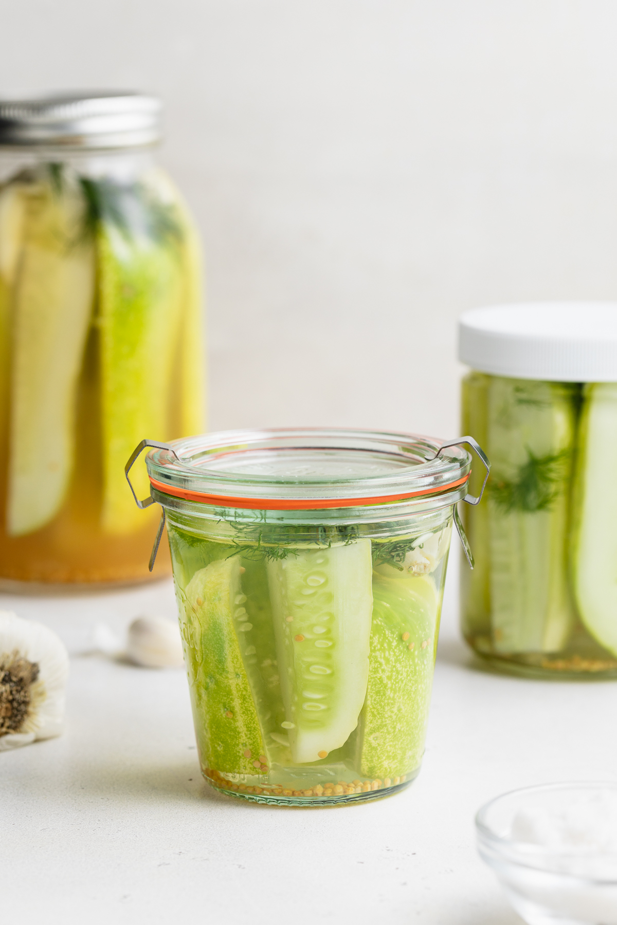 three jars of pickles with garlic and salt on the side.