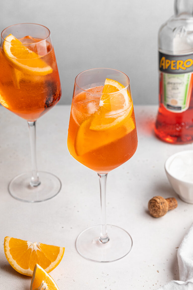 The Perfect Aperol Spritz Two aperol spritz in white wine glasses with a bottle of aperol on the side.