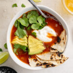 overhead image of Mexican soup with avocado, cilantro, sour cream, and tortillas on top.