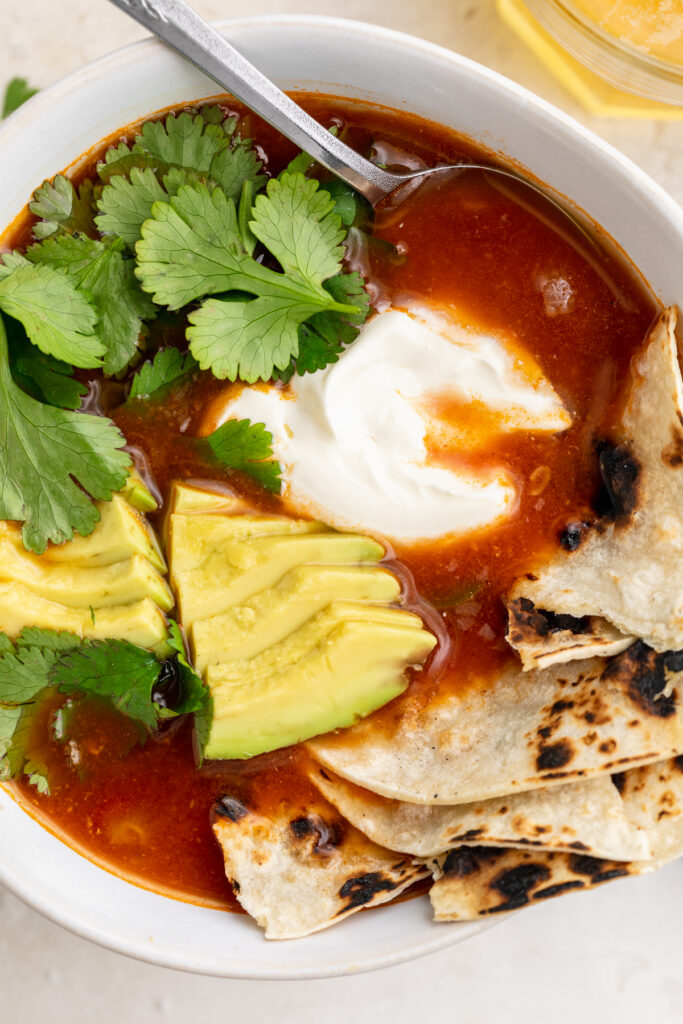 close-up overhead image of Mexican soup with avocado, cilantro, sour cream, and tortillas on top.