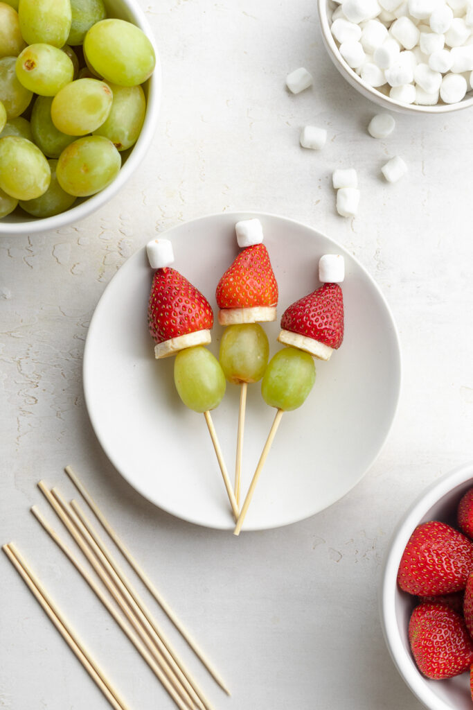 grinch fruit kabobs on a plate with green grapes, strawberries, mini marshmallows, and skewers on the side.